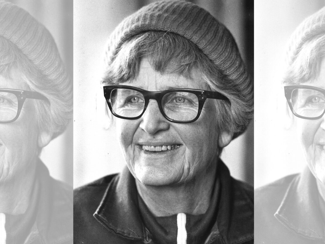 A black and white portrait of Milly. Her head and neck are visible. She is wearing a hat and glasses and she is smiling.