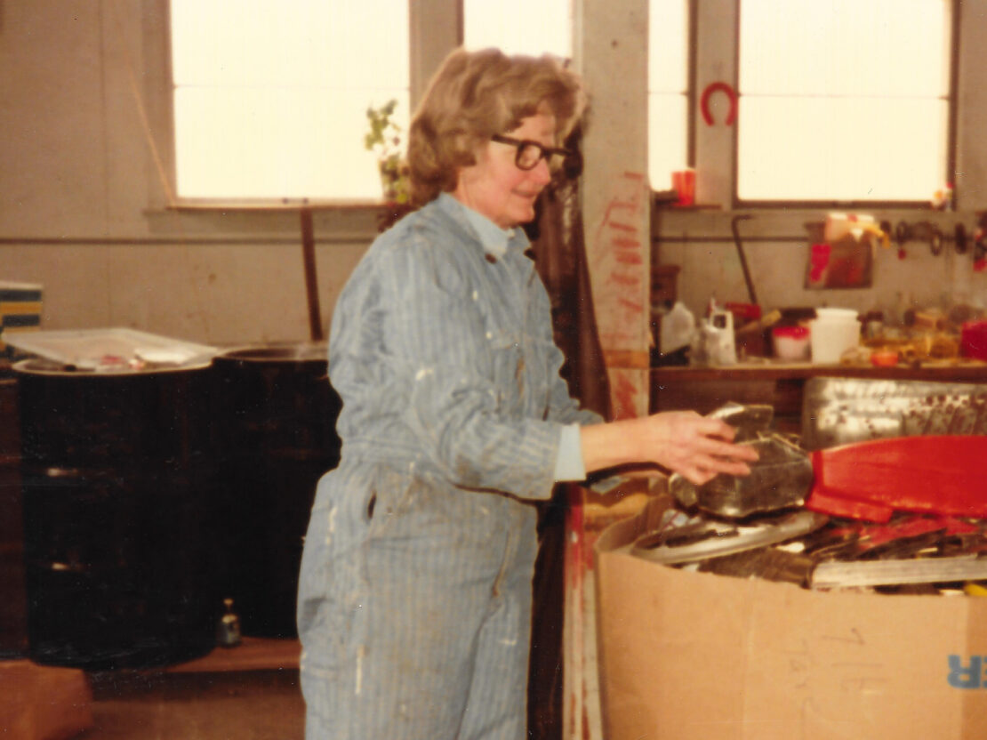 A color photograph showing Milly next to a large cardboard bin that holds lots of items. Milly is turned towards the viewer's right and has her right arm outstretched.