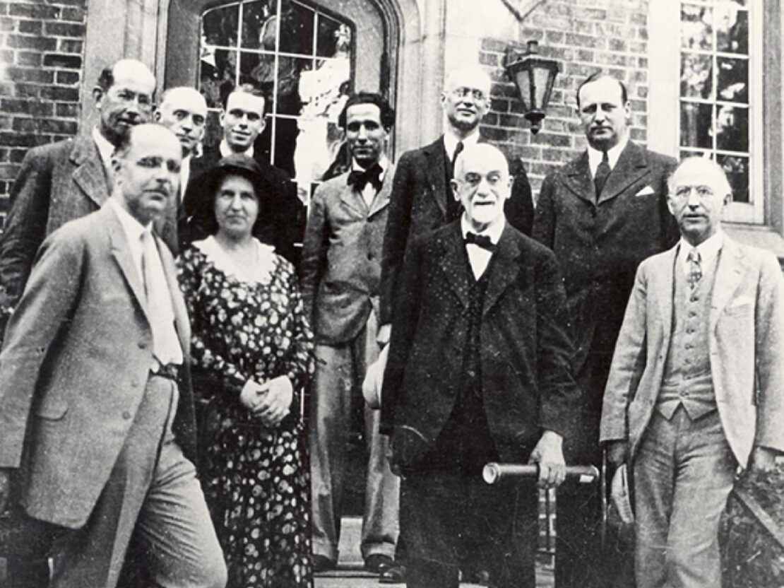 A black and white image of Stephen standing on the steps outside of a brick building with a group of people behind him.