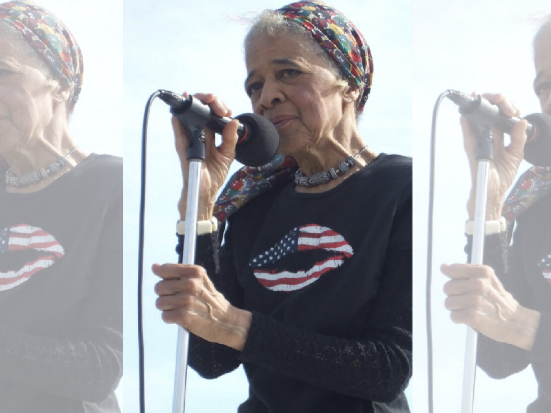 A color photograph of Vel outside holding a microphone and stand and speaking into it.