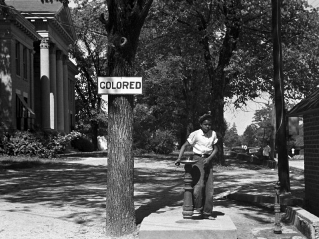 A black and white image of a boy at a drinking fountain outside. On the tree next to the drinking fountain is a sign reading “Coloured.”