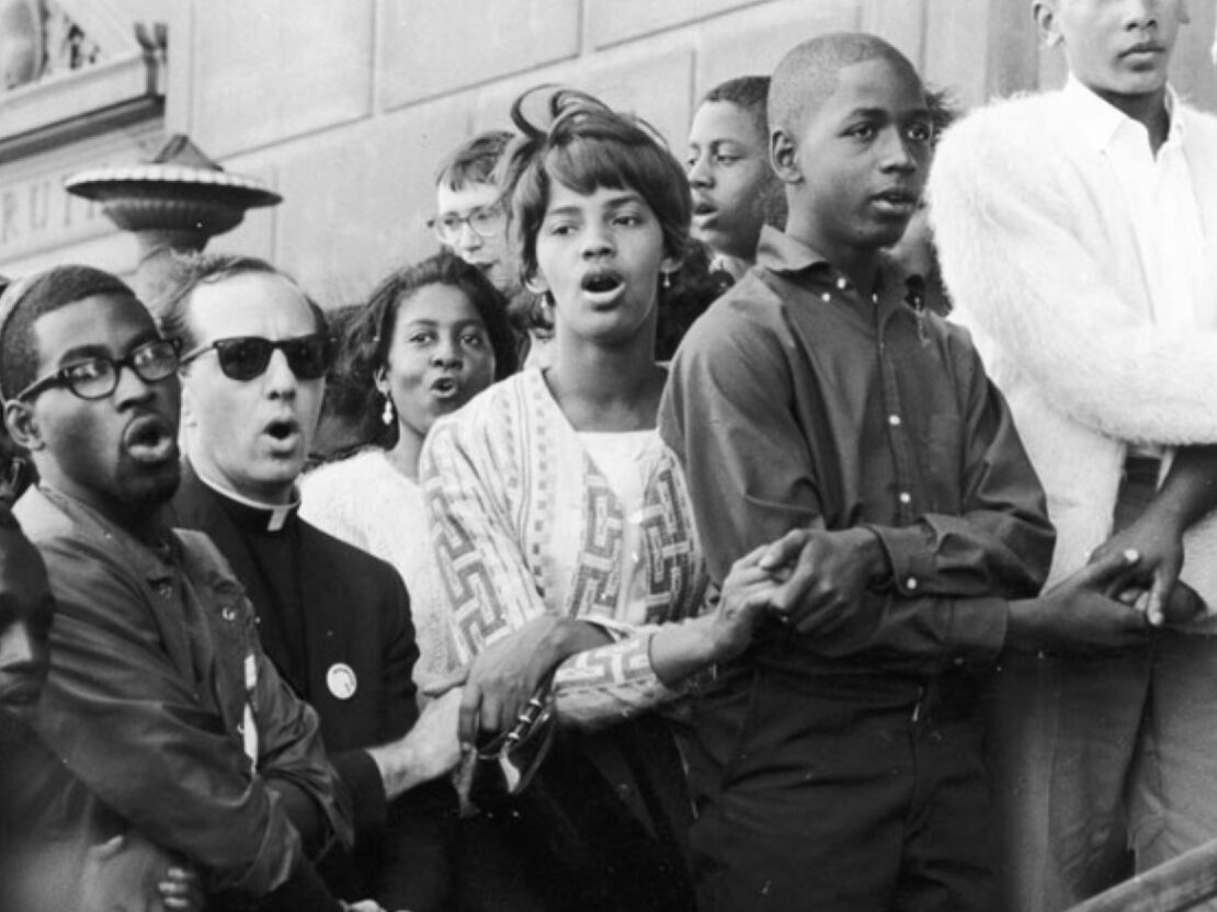 A black and white image of Father Groppi holding hands and singing with a group of people.