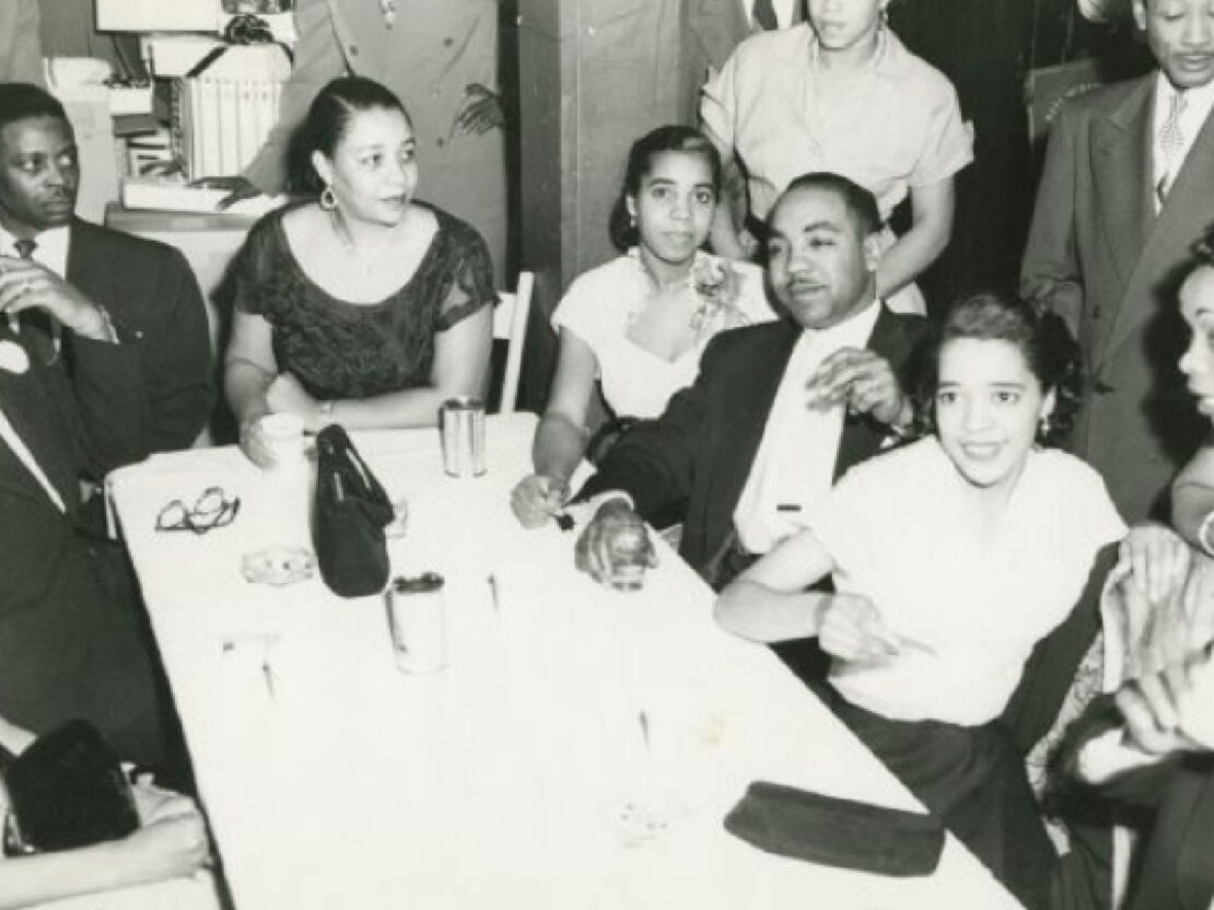 A black and white photograph of Vel seated at a table with many other people. Vel is turned towards the viewer's right, but looks to the viewer's left.