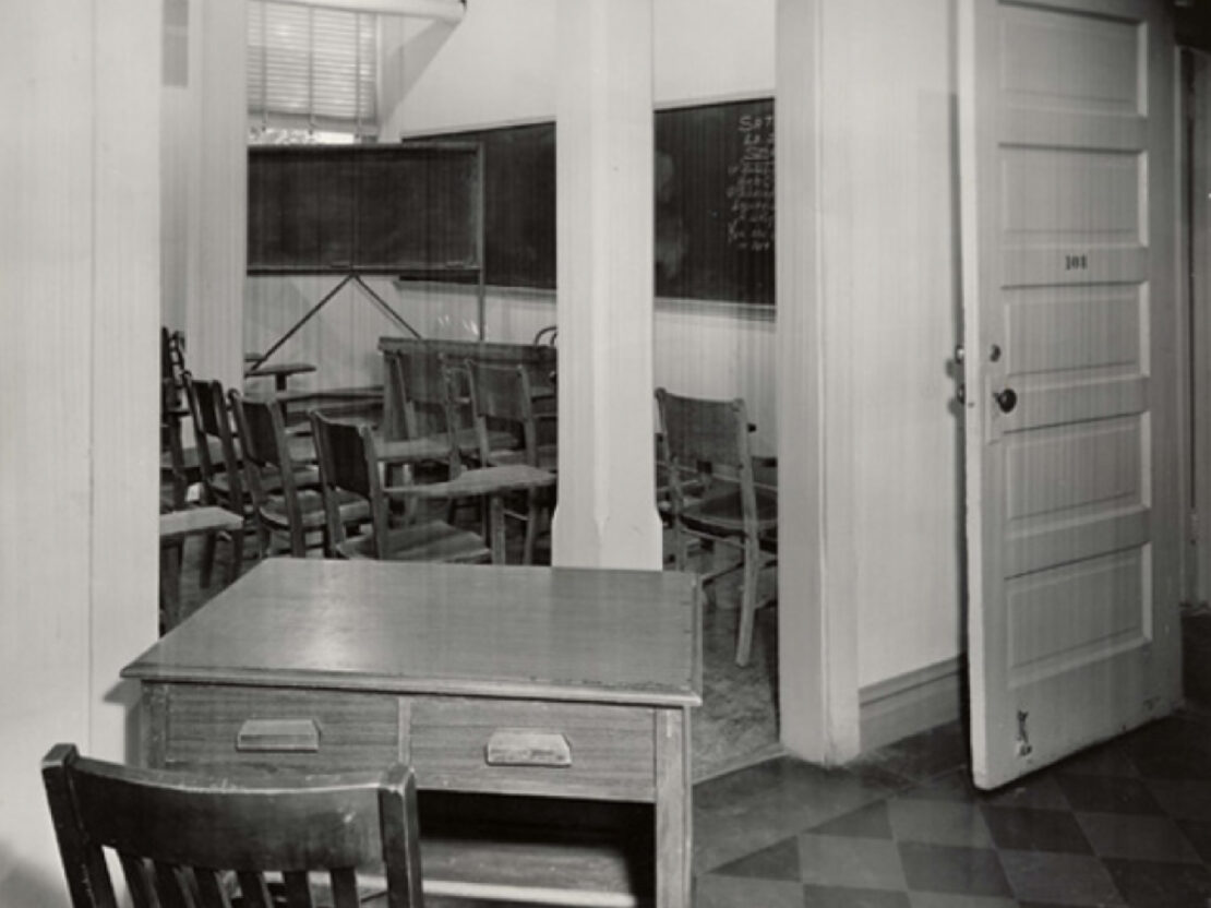 A black and white image of a desk and chair outside of a classroom space with many desks inside it.