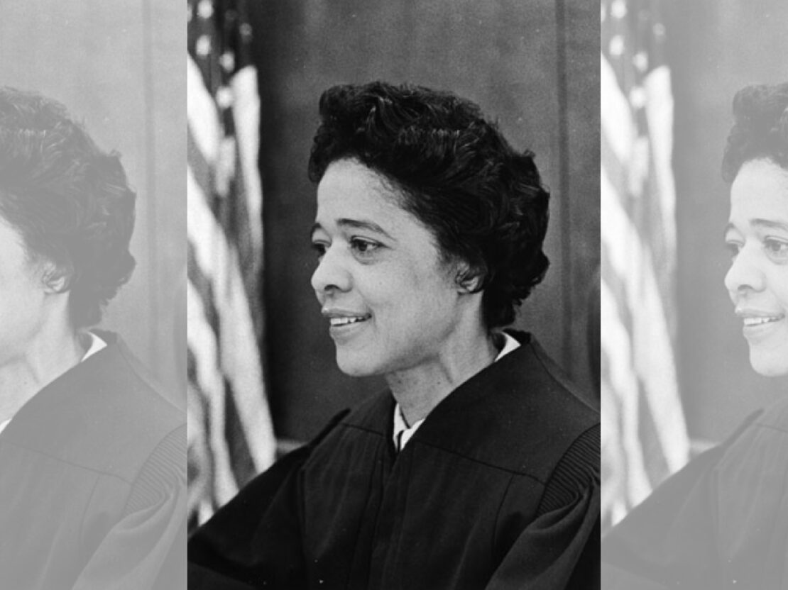 A black and white portrait of Vel wearing judge's robes with a flag visible in the left side of the image.