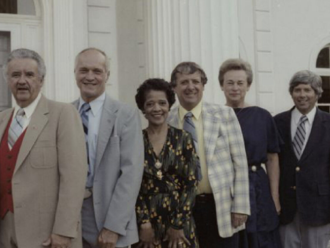 A color photograph showing Vel with the Governor, Lieutenant Governor, and other officials, standing in front of a building.