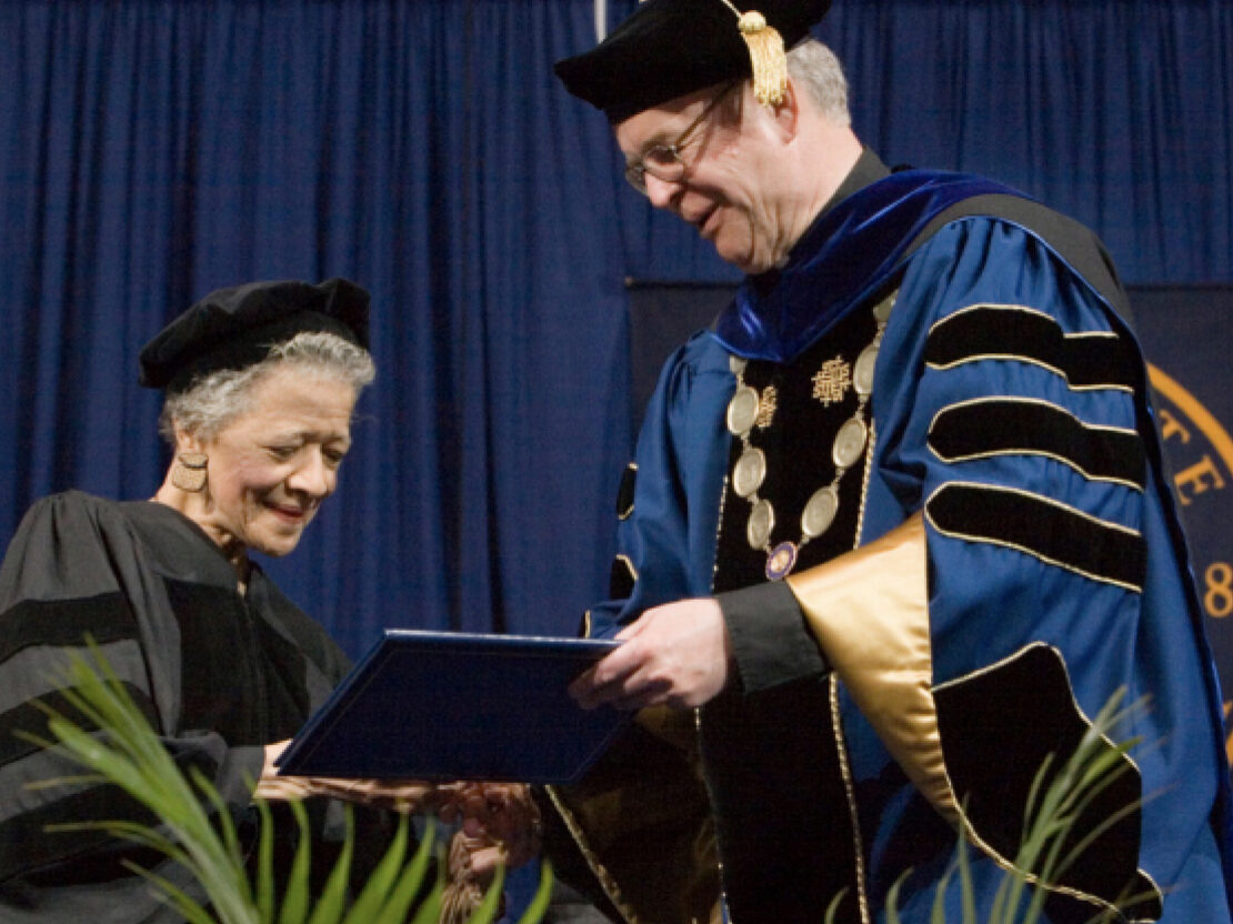 A color photograph showing Vel wearing a cap and gown and holding a diploma being handed to her.