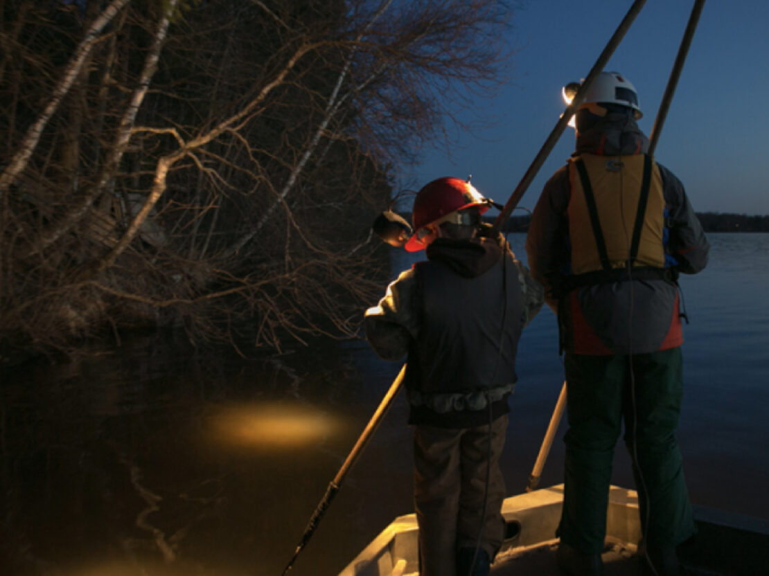 A color still image of two children standing in a boat on the water at night, holding spears and looking down into the water with their headlamps creating spotlights on the water.