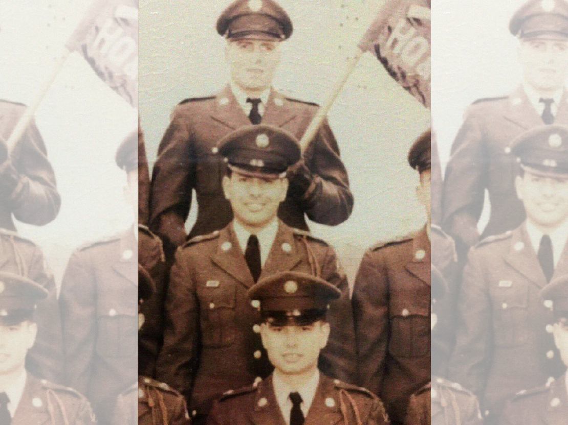 A black and white photograph showing Mahmoud Othman Atta and several other soldiers in the US Army. Everyone is wearing a formal Army uniform and cap. Mahmoud is at the center and is smiling and looking at the camera. Behind him another cadet holds a flag at an angle.