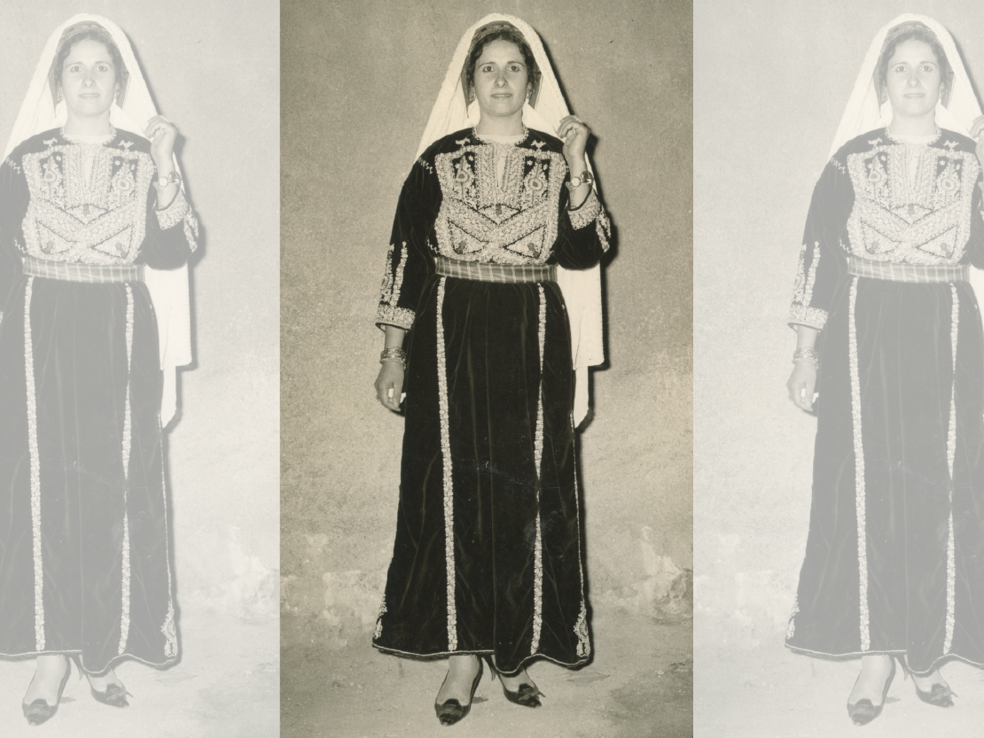 A black and white photograph of a woman in traditional Palestinian dress. She wears a white veil over her head and a dark dress with ornate decoration on the torso and cuffs of the sleeves.