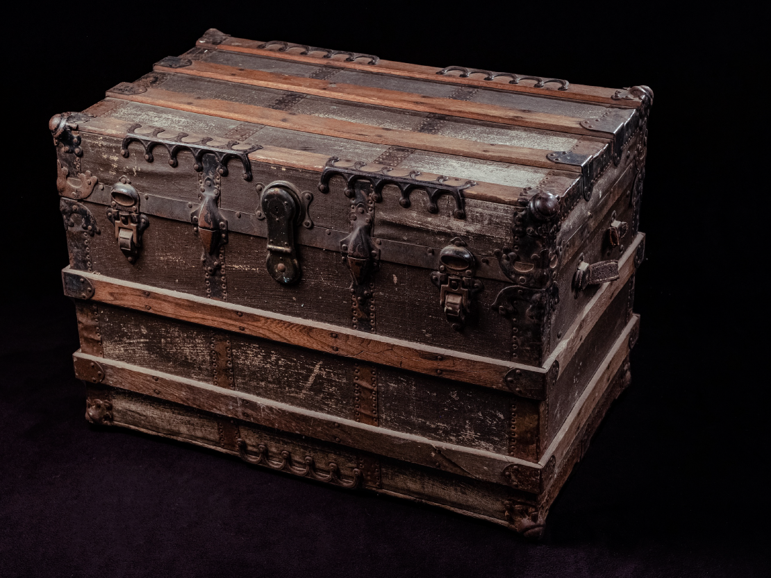 A photograph of a large wooden trunk. The trunk is dark brown and looks scuffed in places due to use and age. There are heavy metal clasps and hinges around the top lid of the trunk.
