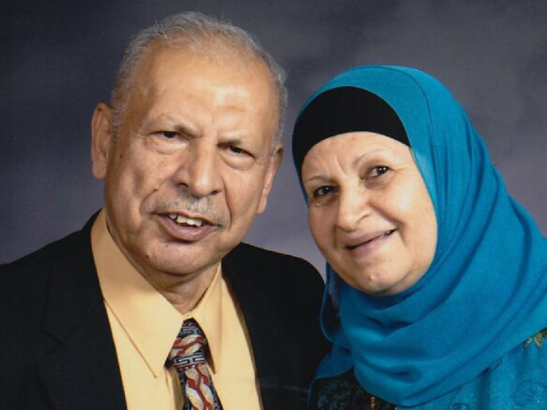 A photograph of Mahmoud and his wife Intisar from when they are older. Mahmoud wears a dark suit jacket, yellow shirt, and patterned tie. Intisar wears a dark blue shirt and lighter blue head scarf. They are both smiling and looking toward the camera.